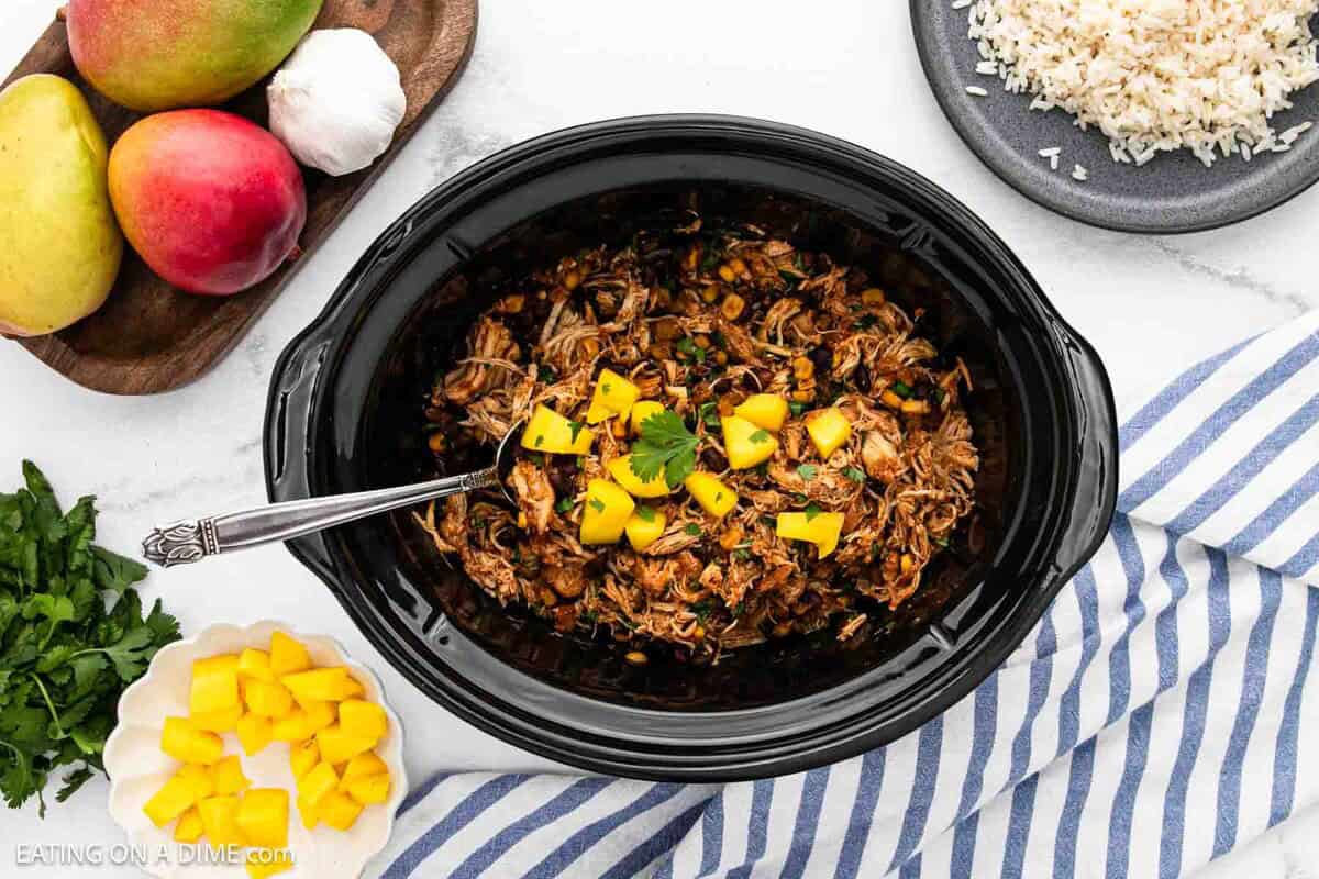 Overhead view of Crockpot Mango Salsa Chicken in a slow cooker, garnished with chopped mango and cilantro. Surrounding the cooker are a plate of mangoes and garlic, a bowl of cooked rice, diced mango, and fresh herbs.