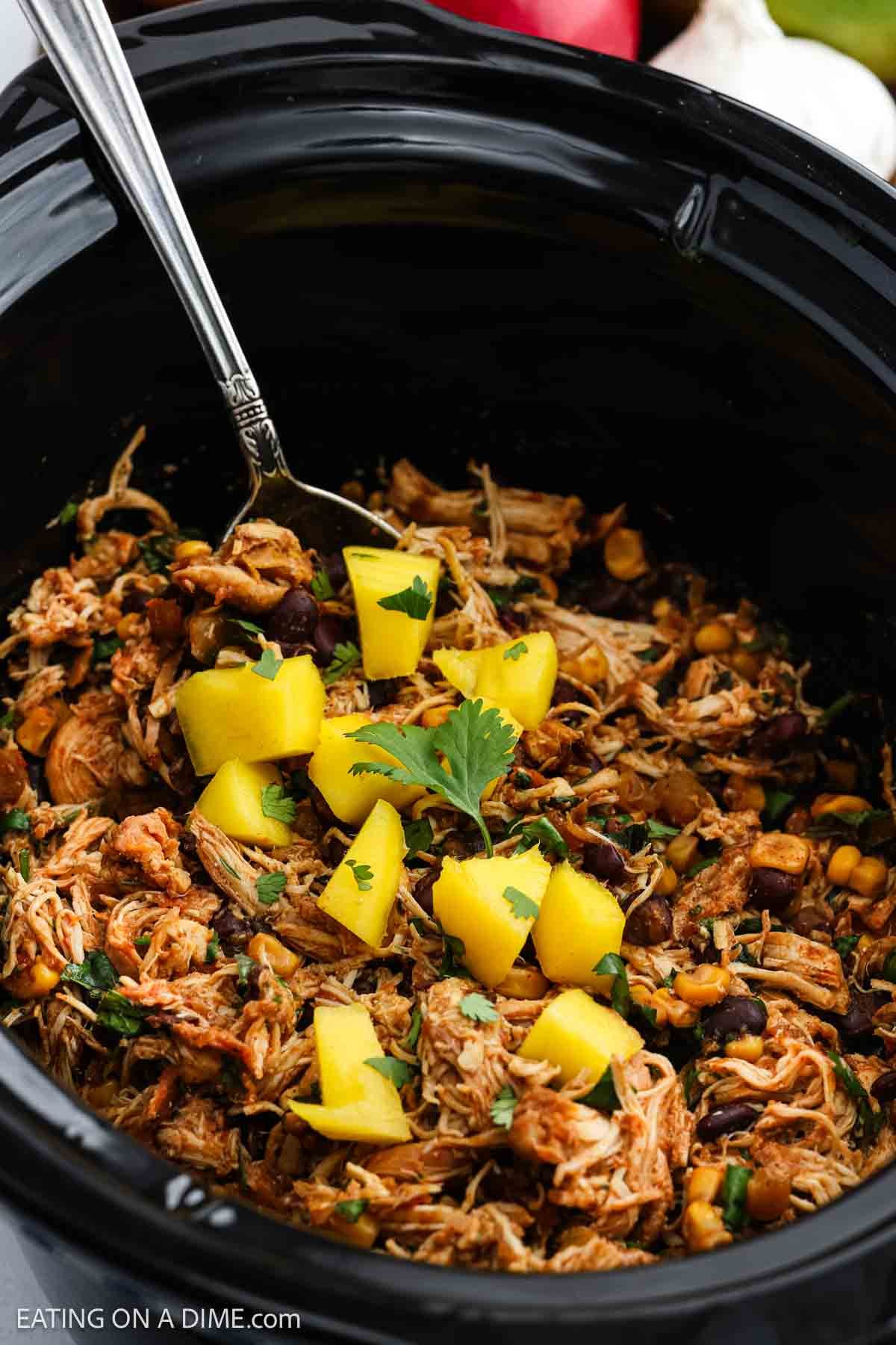 Shredded chicken, black beans, and corn in a black Crockpot Mango Salsa Chicken, topped with mango chunks and cilantro, with a fork resting inside.