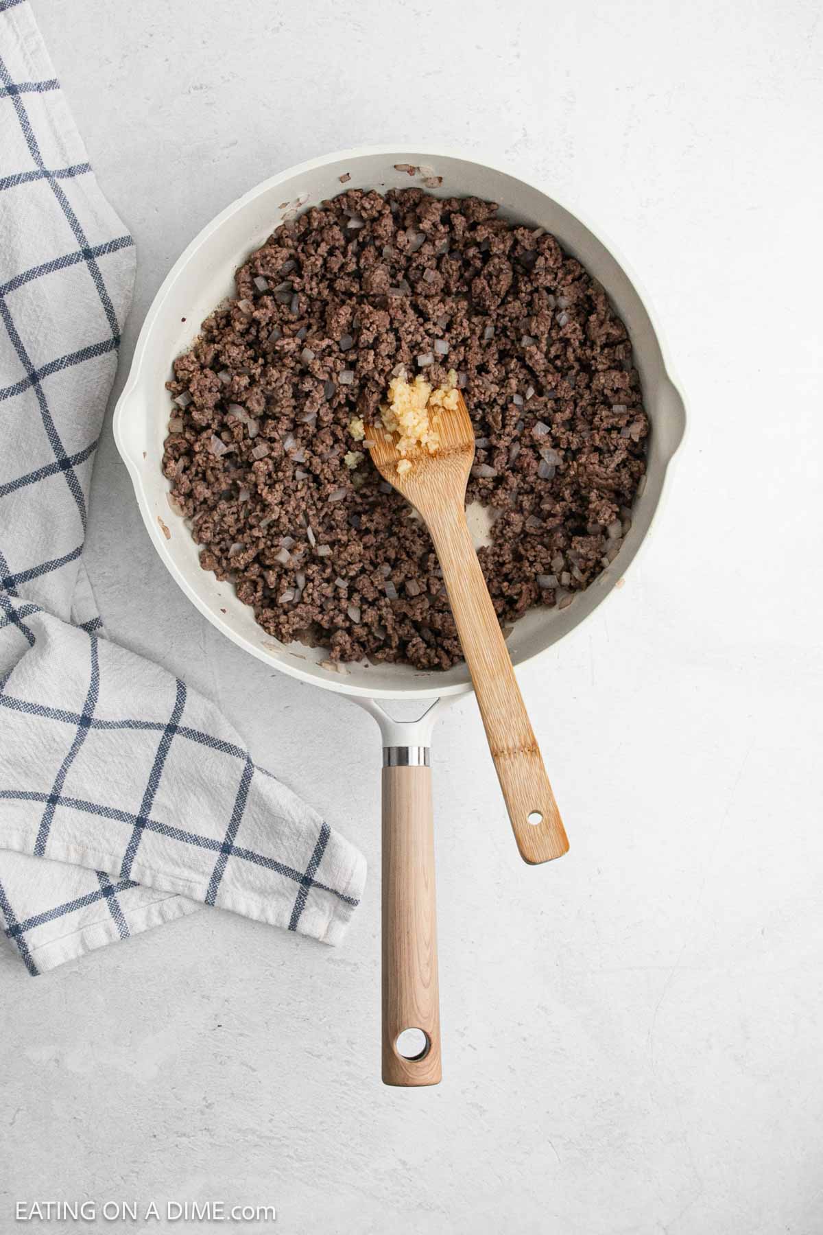 Cooked ground beef in a skillet with minced garlic and a wooden spoon.