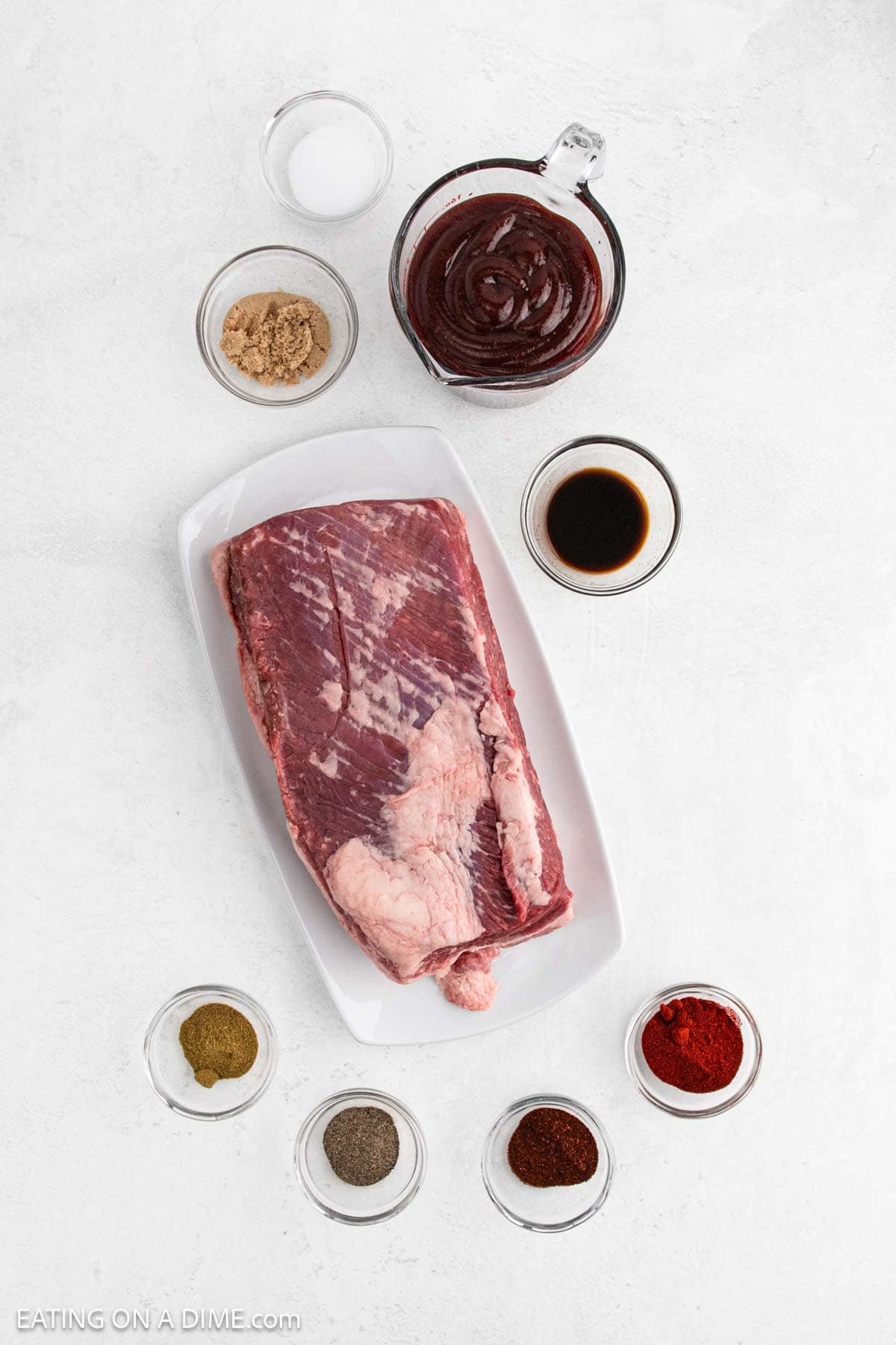 A slab of raw brisket on a white plate, surrounded by small bowls of spices, brown sugar, salt, black pepper, paprika, barbecue sauce, and Worcestershire sauceโperfect ingredients for Crock Pot BBQ Brisketโset on a white background.