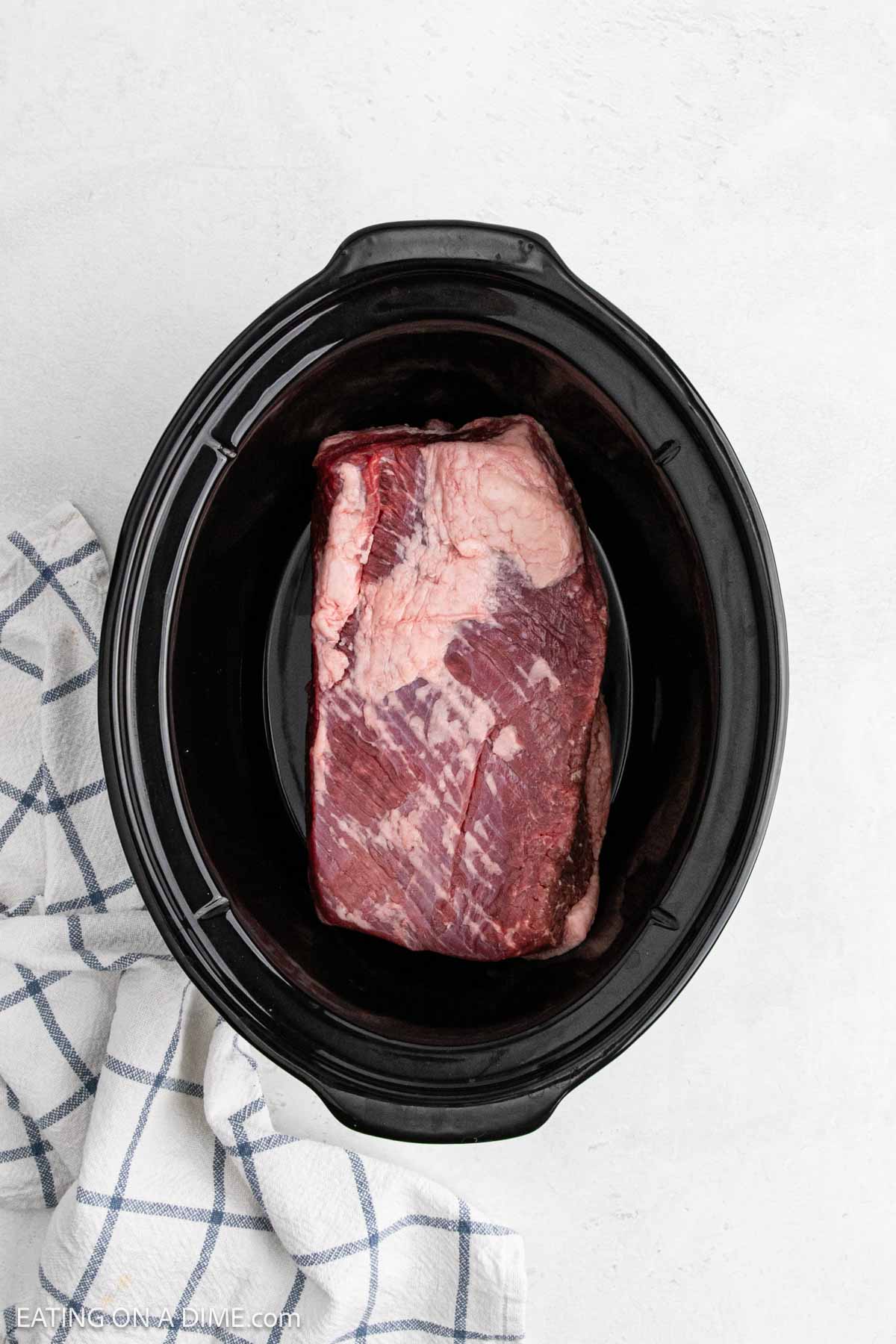 A raw beef brisket sits inside a black slow cooker on a white surface, ready to become delicious Crock Pot BBQ Brisket, next to a folded white and blue checkered kitchen towel.