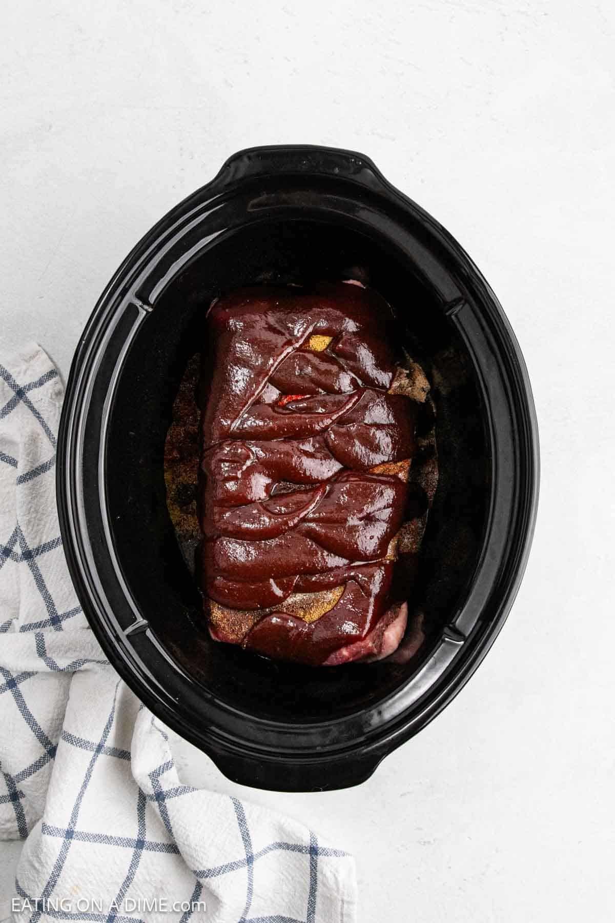 A raw piece of meat coated in barbecue sauce sits inside a black Crock Pot, ready to become tender Crock Pot BBQ Brisket, next to a white and blue checkered kitchen towel on a light background.