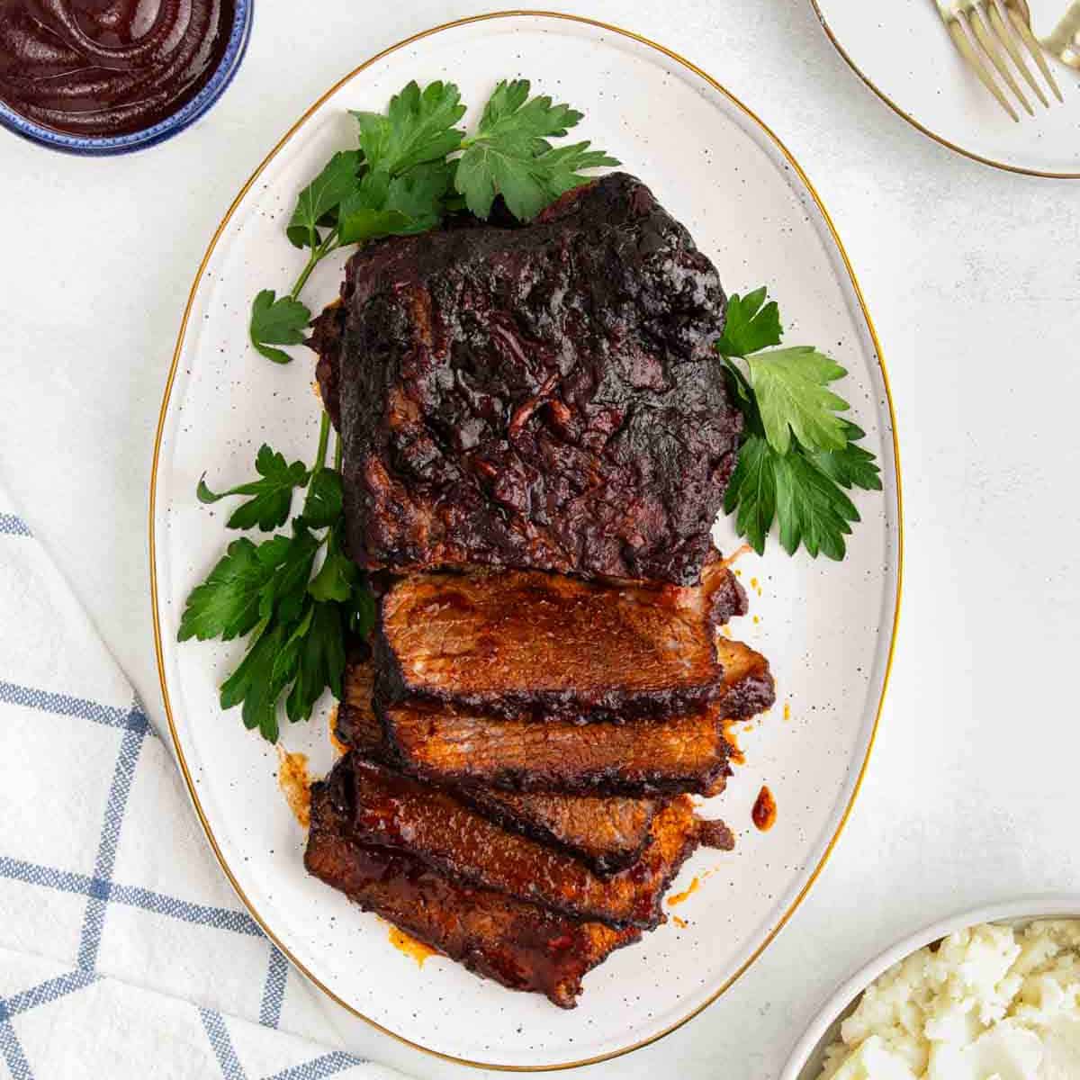 An oval platter with sliced Crock Pot BBQ Brisket, garnished with fresh parsley, sits on a white table next to a bowl of barbecue sauce, creamy mashed potatoes, and a blue-and-white towel.