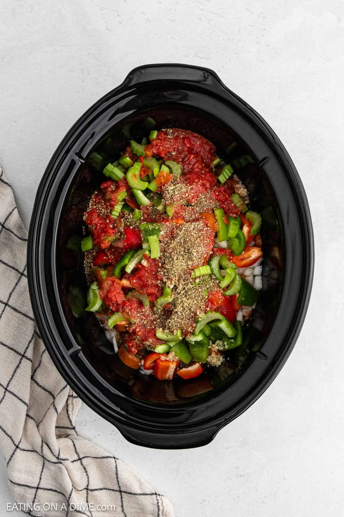 A slow cooker filled with chopped onions, green bell peppers, diced tomatoes, Italian seasoning, and raw sausage for Slow Cooker Chicken Jambalaya sits on a light surface, with a checkered kitchen towel beside it.