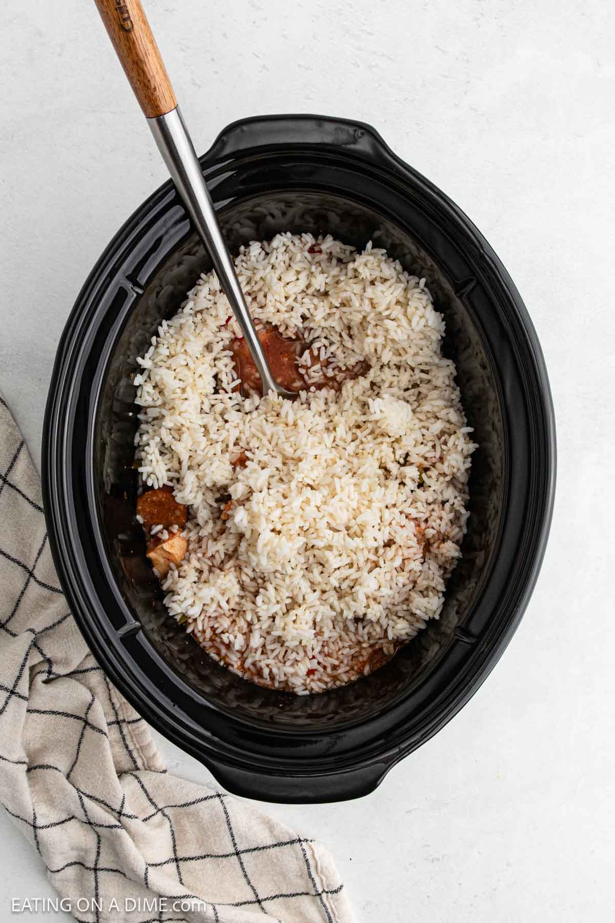A black slow cooker filled with Slow Cooker Chicken Jambalaya—cooked white rice and pieces of meat—being stirred with a wooden spoon. A beige and black checkered kitchen towel rests nearby on a light surface.