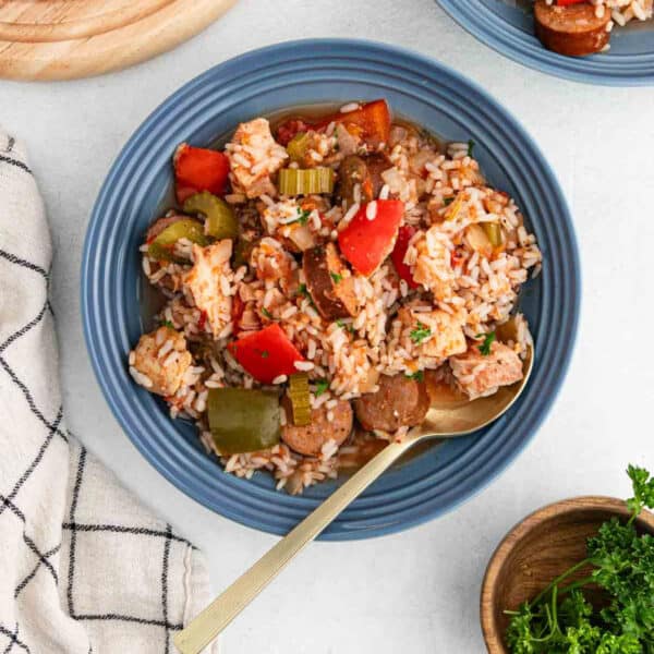A blue bowl filled with Slow Cooker Chicken Jambalaya—rice, sliced sausage, chicken, and colorful bell peppers—with a gold spoon inside. A cloth napkin and a small bowl of parsley sit nearby.
