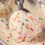 A scoop of creamy, no churn birthday cake ice cream with colorful sprinkles is being lifted from a metal container. The vanilla base delights with vibrant red, green, blue, and orange sprinkles scattered throughout.