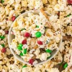 A bowl of Christmas Crunch—a festive mix of popcorn, red and green candies, pretzel sticks, and colorful sprinkles—surrounded by more of this cheerful holiday snack.