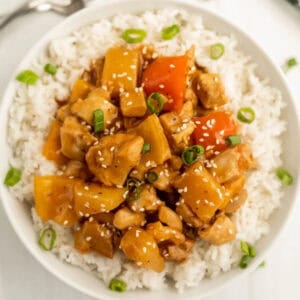 A bowl of white rice topped with Crock Pot Hawaiian Chicken, featuring chunks of chicken, yellow and red bell peppers, pineapple, and garnished with sliced green onions and sesame seeds.