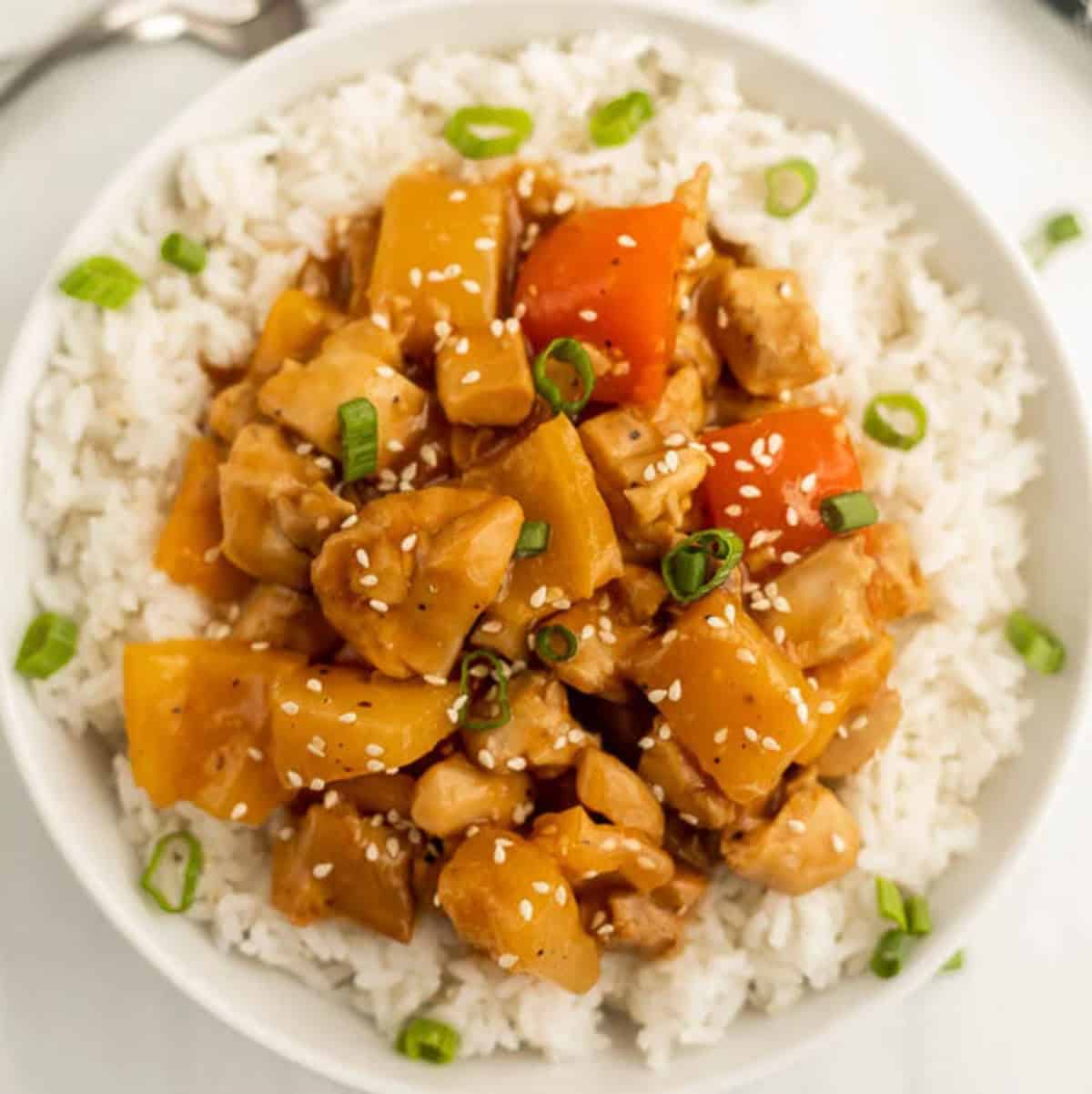 A bowl of white rice topped with Crock Pot Hawaiian Chicken, featuring chunks of chicken, yellow and red bell peppers, pineapple, and garnished with sliced green onions and sesame seeds.