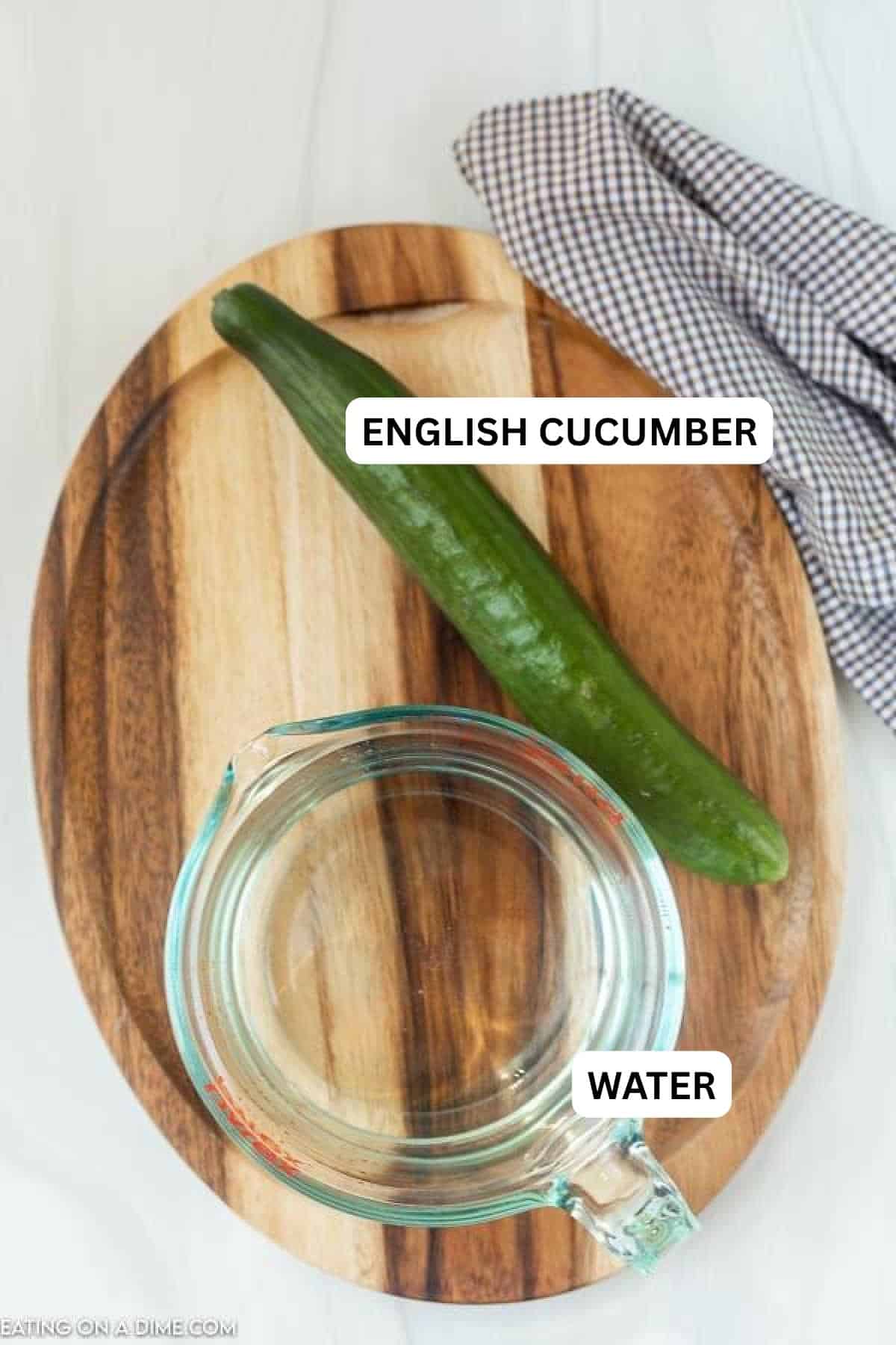 A wooden cutting board holds a fresh cucumber and a glass measuring cup.