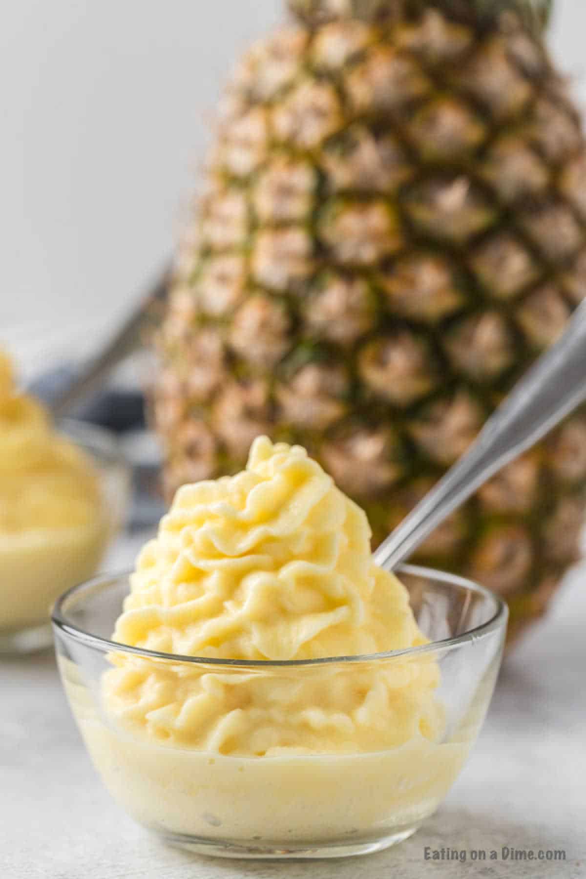 A bowl of creamy, yellow pineapple ice cream reminiscent of the iconic Disney Dole Whip Recipe rests alongside a whole pineapple. The dessert is swirled to perfection, with another partial bowl visible in the background and a spoon ready for indulgence.