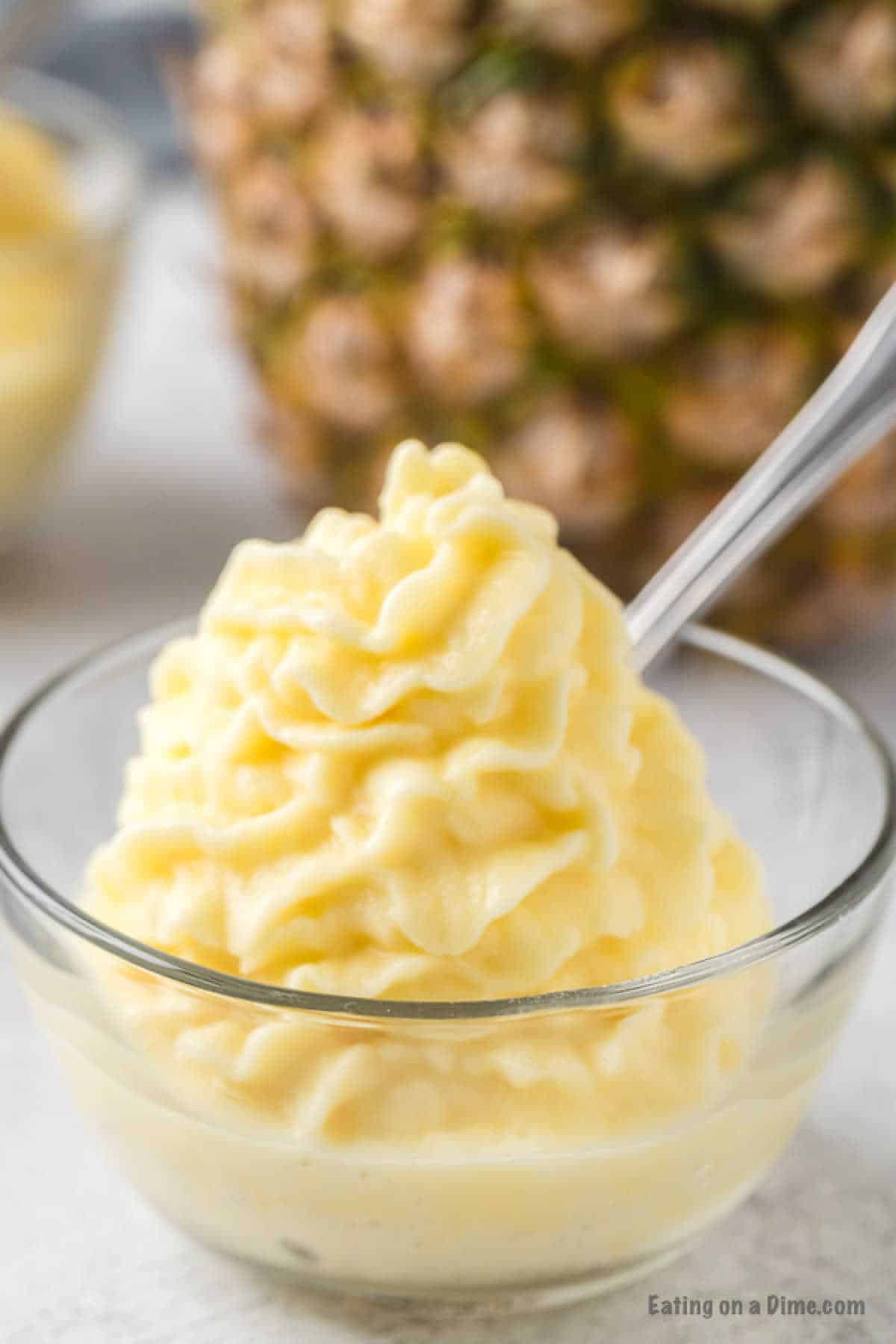 A clear glass bowl filled with creamy, swirled pineapple Dole Whip, reminiscent of the classic Disney Dole Whip recipe. A clear spoon is inserted into the whip. In the background, there is a blurred view of a pineapple.