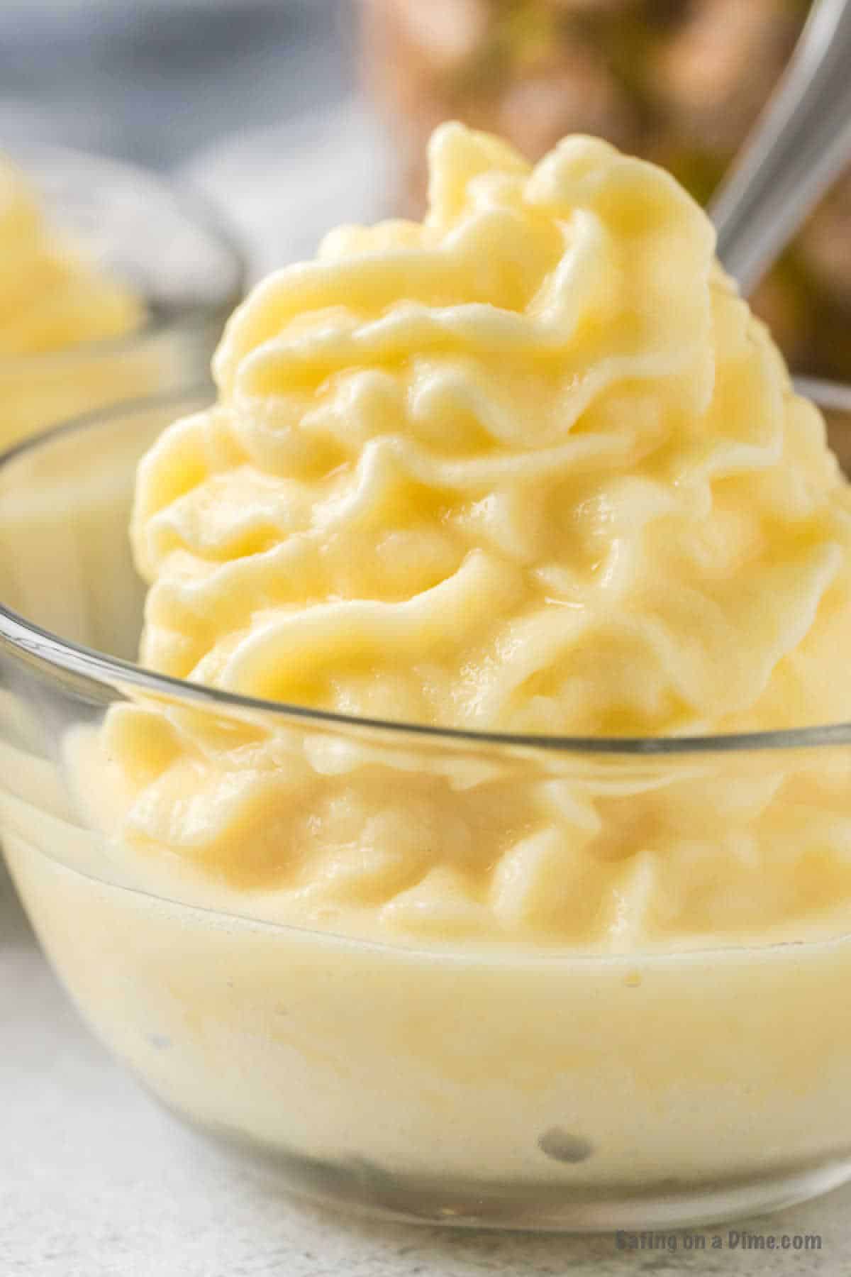 A clear glass bowl filled with creamy, swirled soft serve reminiscent of a Disney Dole Whip. The texture is smooth and slightly fluffy, with a light yellow color from the pineapple flavor. Another dessert can be seen blurred in the background.