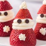 Three Strawberry Santas, decorated with whipped cream faces and hats, and chocolate chip eyes, are arranged on a plate with whipped cream star decorations.