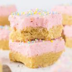 Two stacked Sugar Cookie Bars with thick pink frosting and colorful sprinkles on top. The top bar has a bite taken out, showing the soft texture inside. Other frosted Sugar Cookie Bars are blurred in the background.