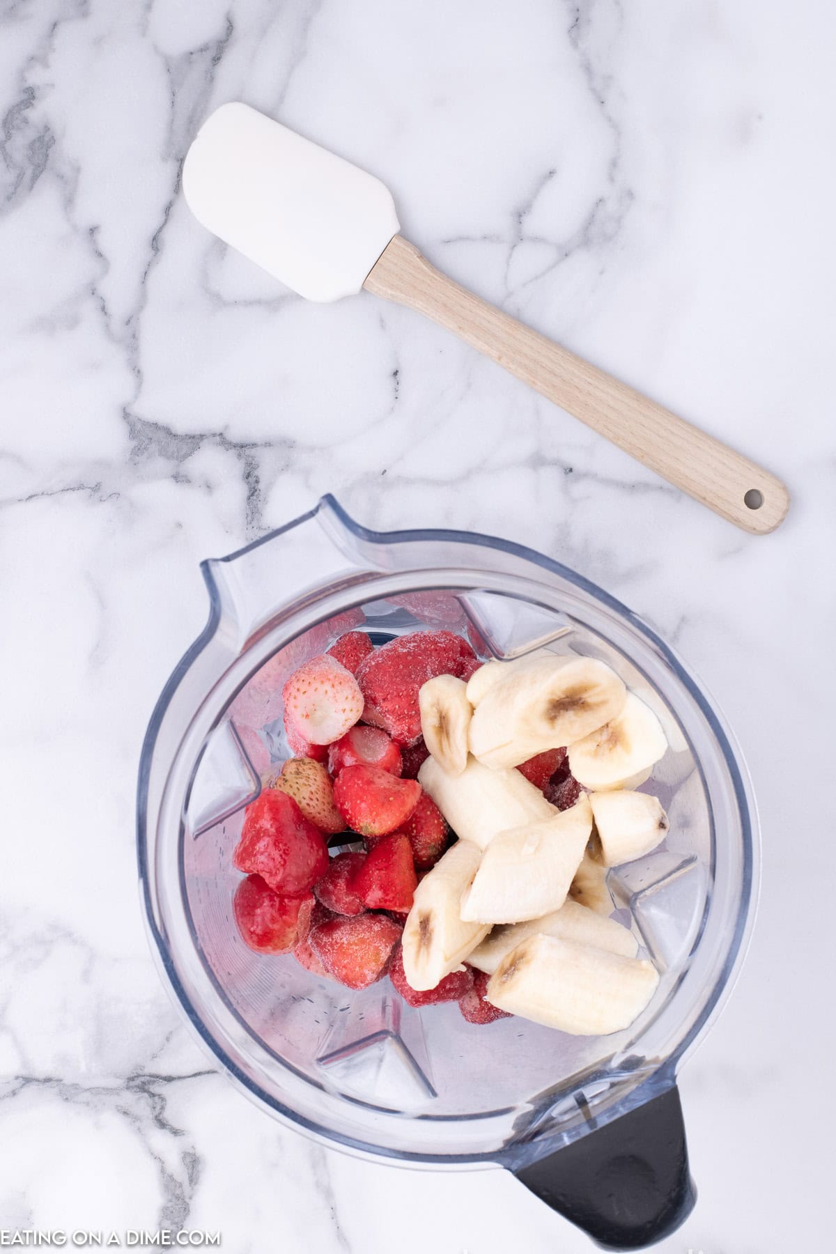 A blender containing sliced bananas and strawberries for homemade fruit ice cream sits on a white marble surface, with a white spatula with a wooden handle placed above it.
