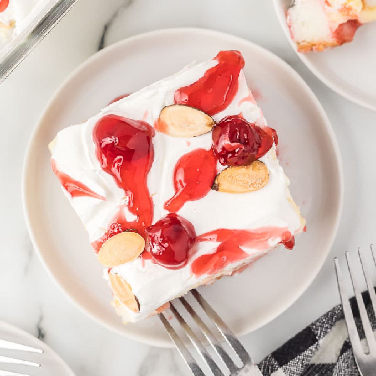 A slice of heaven on earth cake rests on a white plate, adorned with whipped topping, cherry sauce, and sliced almonds. Two forks lie ready on the marble surface beside this delightful dessert.