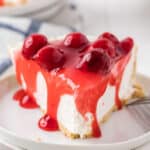 A slice of Philadelphia No Bake Cheesecake with a graham cracker crust topped with glossy cherry pie filling rests on a white plate. A fork is placed invitingly beside the dessert.
