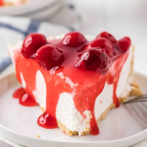 A slice of Philadelphia No Bake Cheesecake with a graham cracker crust topped with glossy cherry pie filling rests on a white plate. A fork is placed invitingly beside the dessert.