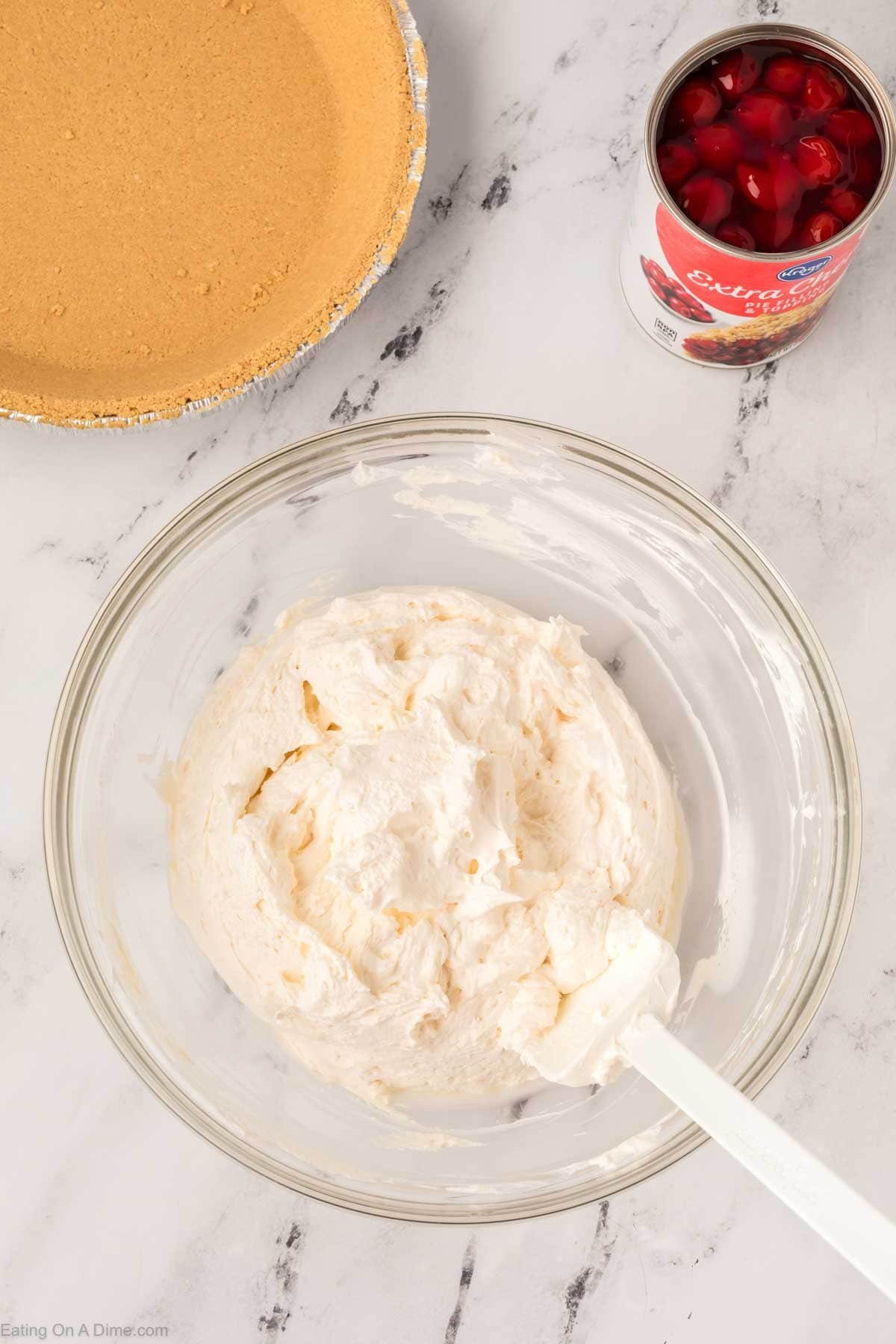 A glass bowl with creamy Philadelphia No Bake Cheesecake filling and a spatula sits on a marble surface, next to a graham cracker pie crust and an open can of cherry pie filling.