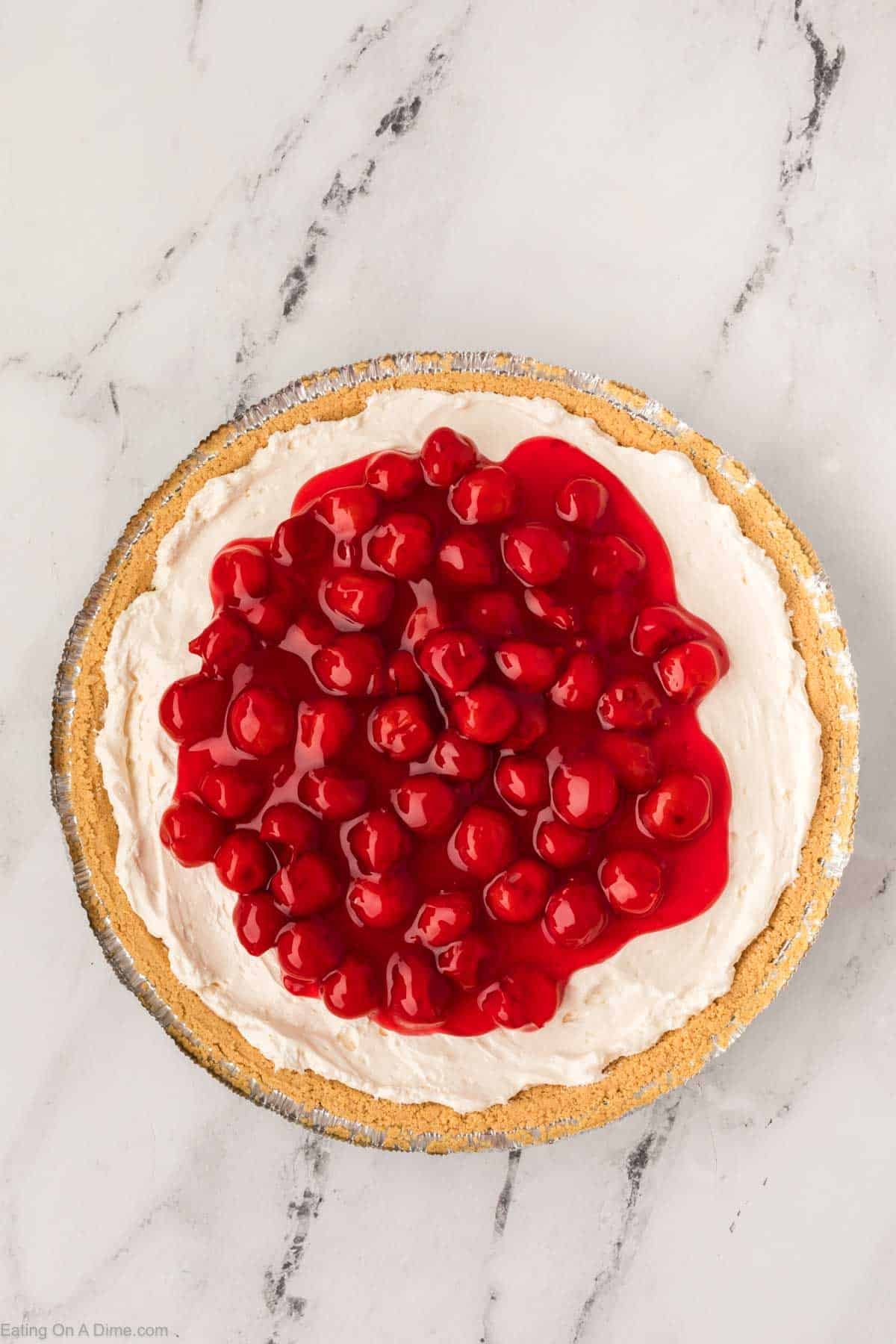 A Philadelphia No Bake Cheesecake pie with a graham cracker crust, creamy filling, and glossy cherry topping, displayed on a white marble surface.