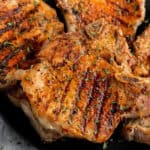 Bone-in pork chops on the grill with perfect char marks, seasoned with herbs, and elegantly served on a sleek black plate.