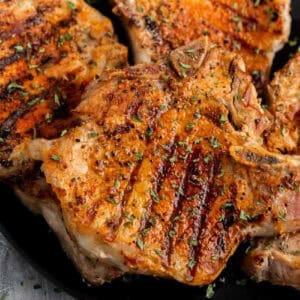 Bone-in pork chops on the grill with perfect char marks, seasoned with herbs, and elegantly served on a sleek black plate.