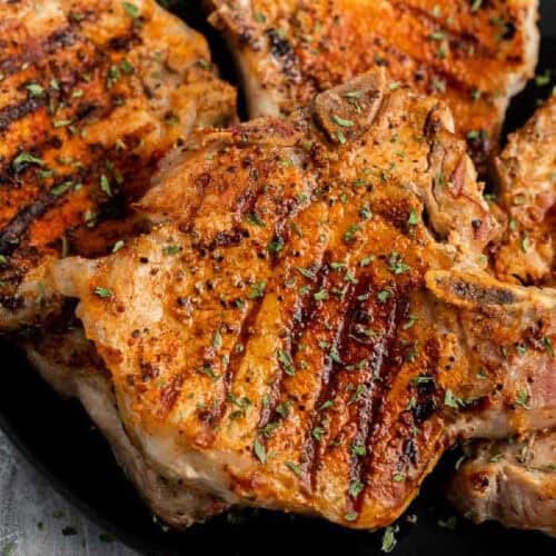 Bone-in pork chops on the grill with perfect char marks, seasoned with herbs, and elegantly served on a sleek black plate.