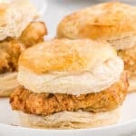 A close-up of a Chick-Fil-A Chicken Biscuit sandwich filled with crispy fried chicken, with two more chicken biscuit sandwiches blurred in the background.