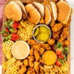 A wooden platter, reminiscent of a Chick-Fil-A Charcuterie Board, boasts chicken sandwiches, waffle fries, crispy fried chicken pieces, strawberries, pickles, and two savory dipping sauces in small bowls. A red and white checkered cloth is artfully draped on the side.