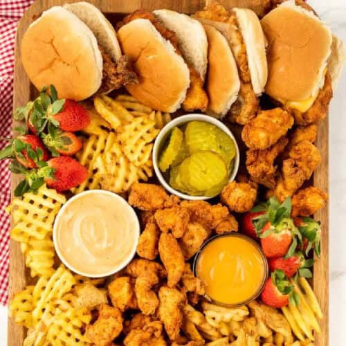 A wooden platter, reminiscent of a Chick-Fil-A Charcuterie Board, boasts chicken sandwiches, waffle fries, crispy fried chicken pieces, strawberries, pickles, and two savory dipping sauces in small bowls. A red and white checkered cloth is artfully draped on the side.