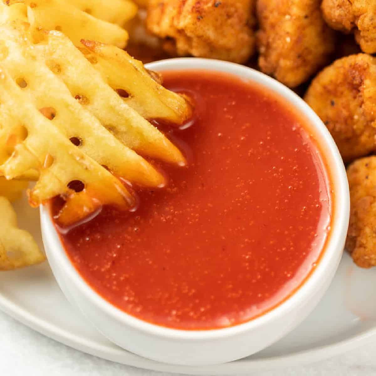A white bowl brimming with Chick-Fil-A Polynesian Sauce is set on a plate. A waffle fry dives into the sauce, surrounded by crispy fried chicken pieces and more fries in the background.
