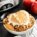 A bowl of Crock Pot Apple Crisp topped with vanilla ice cream sits on a striped cloth. In the background, red apples and part of a slow cooker add a cozy touch.