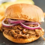 A close-up of a pulled pork sandwich on a bun, topped with sliced red onions. The shredded pork looks moist and saucy—perfect inspiration for your next Crock Pot Brisket Sandwich Recipe.
