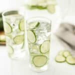 Two tall glasses brimming with cucumber water, ice, and crisp cucumber slices sit on a white surface. A striped cloth and extra slices adorn the scene, crafting an inviting and refreshing tableau.
