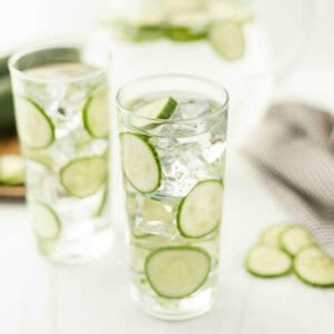 Two tall glasses brimming with cucumber water, ice, and crisp cucumber slices sit on a white surface. A striped cloth and extra slices adorn the scene, crafting an inviting and refreshing tableau.