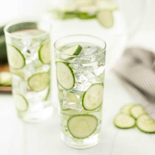 Two tall glasses brimming with cucumber water, ice, and crisp cucumber slices sit on a white surface. A striped cloth and extra slices adorn the scene, crafting an inviting and refreshing tableau.