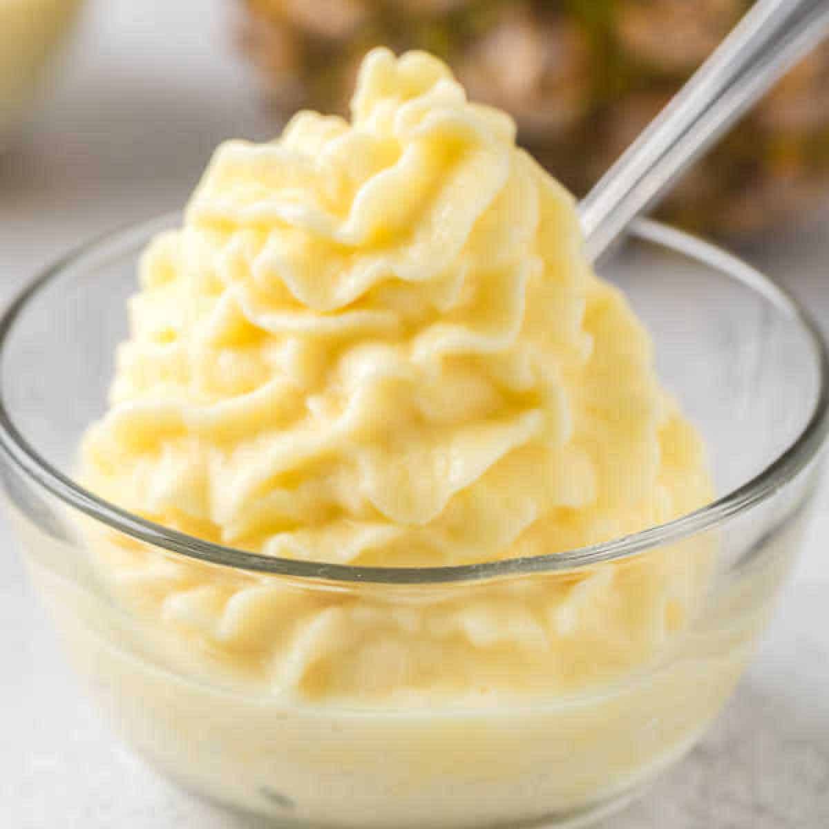 A glass bowl filled with creamy, swirled pineapple soft serve resembling a Disney Dole Whip recipe. A spoon dips into the luscious treat, with a blurred pineapple adding a tropical vibe in the background.