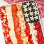 An American Flag Charcuterie Board features cheese stars, blueberries, raspberries, marshmallows, pita chips, red bell peppers, pepperoni, cheese slices, and chopped strawberries artfully arranged on a tray.