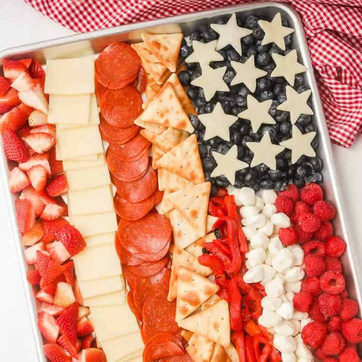 An American Flag Charcuterie Board features cheese stars, blueberries, raspberries, marshmallows, pita chips, red bell peppers, pepperoni, cheese slices, and chopped strawberries artfully arranged on a tray.