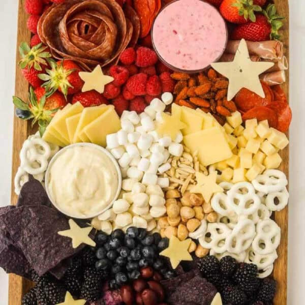A vibrant Red, White, and Blue charcuterie board featuring sliced meats, cheese, raspberries, strawberries, blueberries, marcona almonds, purple tortilla chips, yogurt-covered pretzels, marshmallows, and star-shaped cheese pieces. Two dips are included for added delight.