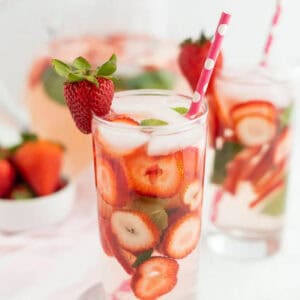 A glass of refreshing Strawberry Water with ice, fresh strawberry slices, mint leaves, and a pink polka dot straw. Another glass and a bowl of strawberries can be seen in the background.