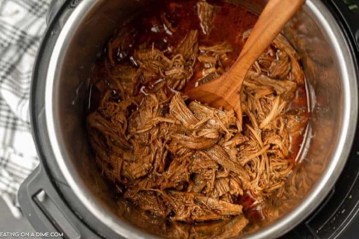 Instant Pot Brisket simmers in a rich sauce, with tender shredded beef being stirred by a wooden spoon. A checkered kitchen towel lies partially visible next to the pot.