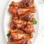 A white oval platter presents eight roasted chicken drumsticks, perfectly glazed with a shiny barbecue sauce. Cooked to perfection in an instant pot, these drumsticks are garnished with fresh parsley sprigs, adding a vibrant pop of green to the dish.