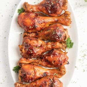 A white oval platter presents eight roasted chicken drumsticks, perfectly glazed with a shiny barbecue sauce. Cooked to perfection in an instant pot, these drumsticks are garnished with fresh parsley sprigs, adding a vibrant pop of green to the dish.