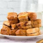 A plate stacked with thick slices of Nutella French Toast, some dusted in cinnamon sugar and others revealing gooey chocolate or cinnamon filling, sits on a white marble surface with a glass of milk in the background.