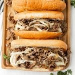 Three Philly cheesesteaks filled with sliced beef, melted cheese, and sautéed onions are arranged on a wooden tray, with fresh parsley scattered nearby.