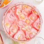 A large glass bowl filled with pink, foamy rainbow sherbet punch is topped with scoops of colorful sherbet. A glass and a striped napkin are nearby.
