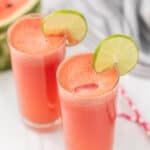 two glasses of watermelon water topped with lime rounds, pink straws and watermelon blurred in the background.